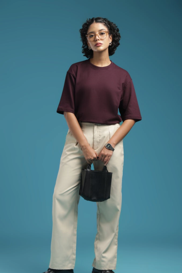 ADEL & VIDA burgundy oversized t-shirt worn by a model with beige trousers and glasses against a blue background.