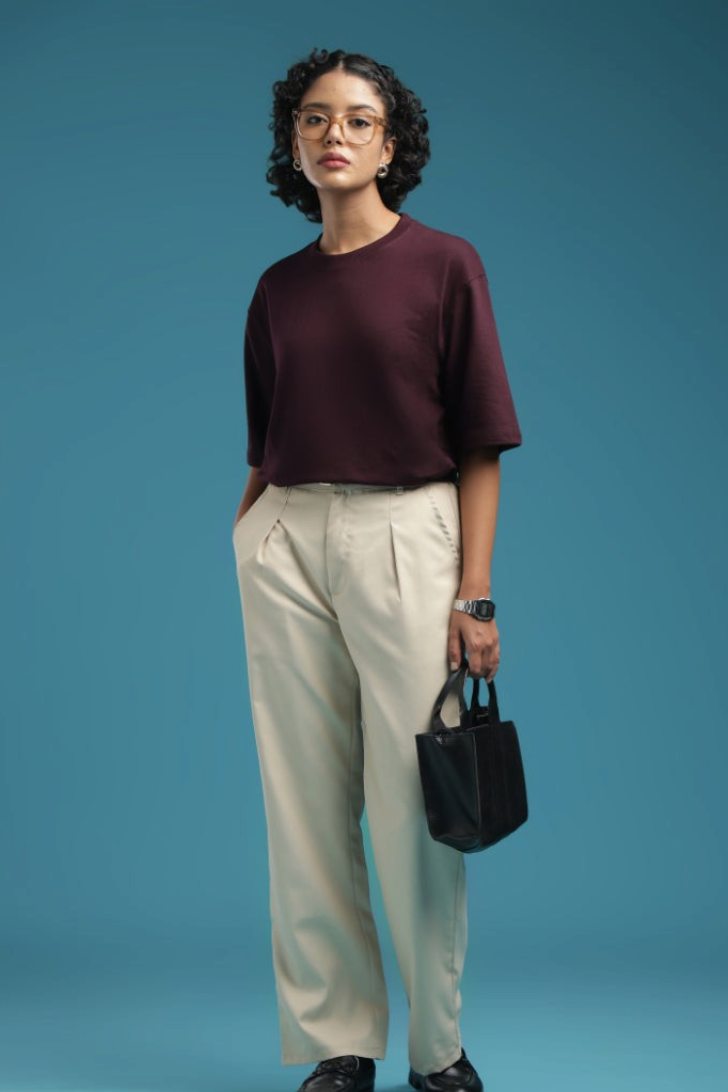 ADEL & VIDA model wearing an oversized burgundy t-shirt with cream trousers and glasses against a blue background